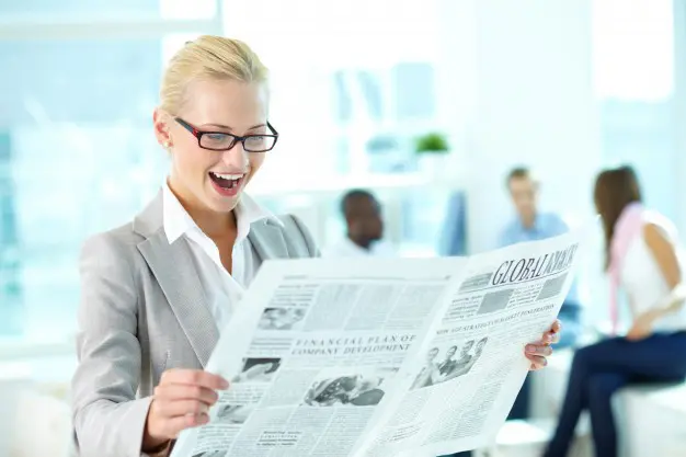 A woman in grey jacket holding newspaper near wall.